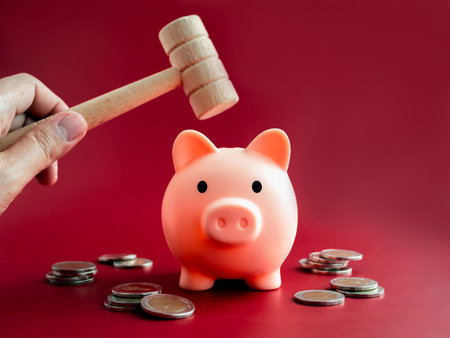 The pink piggy bank, front view is about to be smashed with a wooden hammer in hand over the coins piles on red background.の写真素材