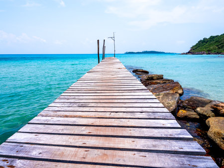 Wooden bridge heading to the blue sea. Brown wood plank pathway bridge on the beach sand with rocks at the local port in the island on sunny day. Seascape summer holiday background.の写真素材