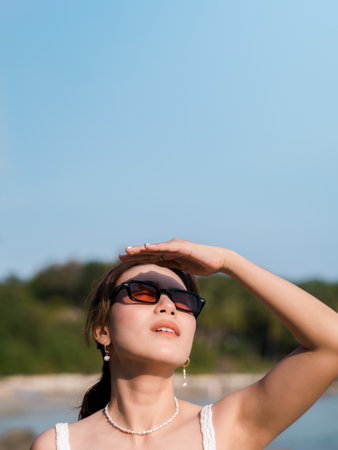 Beautiful Asian woman with sunglasses on the beach in summer vibes with copy space. Attractive female covering the sunlight with hand at the sea and sky background, Holiday vacation, vertical style.の写真素材