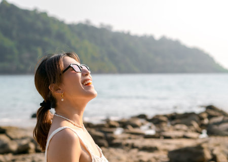 Beautiful Asian woman portrait with sunglasses on the beach in summer vibes. Happy female smile with teeth looking up to sky on sea, island, rocks and sky background, Holiday vacation, summertime.の写真素材