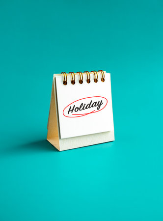 Holiday summer and vacation concept. Relaxing calligraphy font of "Holiday", handwritten and red round mark on small standing desk calendar page on aqua blue background, vertical and minimal style.の写真素材