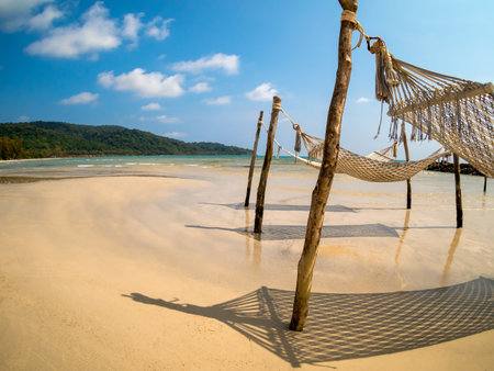 Three empty rope cradle hanging on the wooden poles on sandy beach, island and blue sky background, fisheye view. Beautiful shadow of hanging cradle or swing beds on clean sand beach.の写真素材