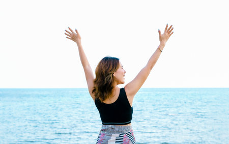 Back of Asian woman in black tank top closing eyes with happy raise hands up pose on the beach in summer vibes. Happy female standing in front of the sea and blue sky background, Holiday vacation.の写真素材