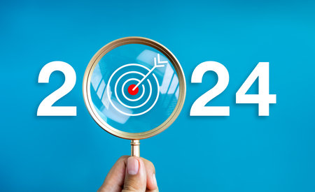 Happy new year 2024 with business concept banner. The big white 2024 year number with target dart icon inside the golden magnifying glass in hand on blue background. Trends, plan and goals concepts.の写真素材
