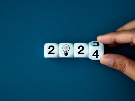 2024 happy new year with creative idea trend concepts. 2023 to 2024 year numbers calendar with target dart and bulb icon on white cube blocks flipping by human hand isolated on blue banner background.の写真素材