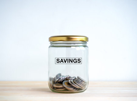 Saving, income, profit, retirement money and financial concept. Word "SAVINGS" on closed glass bottle with gold cap with many coins inside with empty space, on wooden table and white background.の写真素材