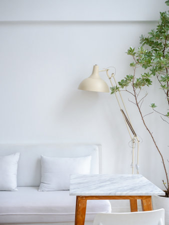 Modern relaxing and clean living room corner decoration with white couch or sofa seat with pillows, beige table light lamp near empty marble top table and green leaves, houseplant, vertical style.の写真素材