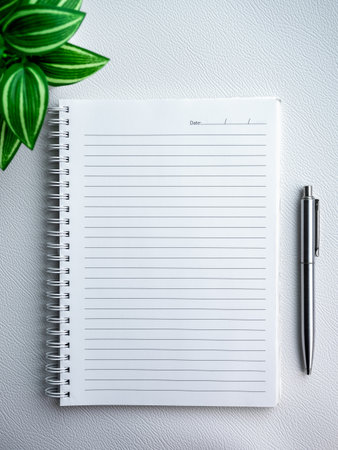 Empty blank of notebook diary page. Template white paper sheet with line and date for note on vertical spiral notepad near a silver pen and green plant isolated on white luxury leather background.の写真素材