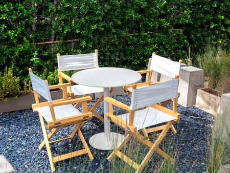 Four empty director chairs, fabric and wood materials with round shape table at outdoor garden on blue gravel stone floor on green tree, bush background. Table set relaxing loft style in the garden.の写真素材