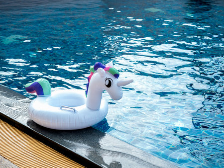 Photo of the white inflatable ring, cute unicorn doll in the outdoor swimming pool with copy space. Empty swim ring for children with sunshine for summer beach.の写真素材