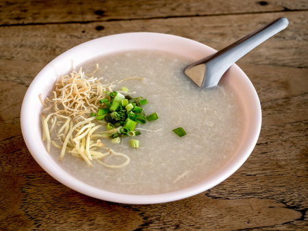 Image of green fresh sliced spring onion and ginger slices on rice porridge in light pink bowl with metal short spoon on wooden table, close up. Asian style cuisine, hot breakfast traditional food.の写真素材