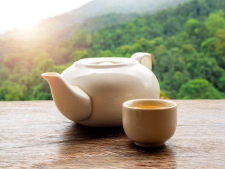 Teapots set. White ceramic teapot with hot tea water in small cup preparing on wooden table on natural green mountain view, countryside background with sunlight in the morning.の写真素材