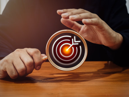 Target, red bullseye dartboard icon inside magnifying glass in businessman hand for focus business goals and trends on wood table. Business marketing strategy, leadership concept, minimal style.の写真素材