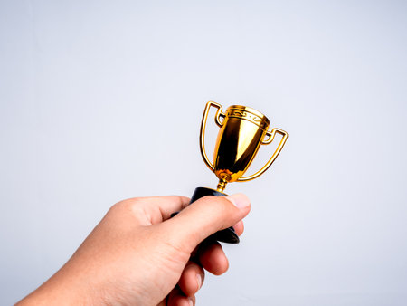 A gold trophy, brass steel, dual handle, in human hand, isolated on white background with copy space. Golden trophy on desk, minimal. Small success, award, reward of effort and winner prize concept.の写真素材