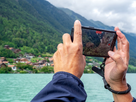 Tourist's hands holding smartphone, taking photo or video of village on green mountain near lake on...