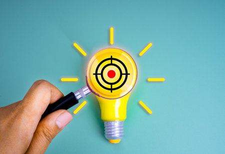 Gun target icon in magnifying glass lens hold by hand on yellow electric light bulb on blue background, minimal style. Goal and success, creativity, inspiration idea, business marketing concepts.の写真素材