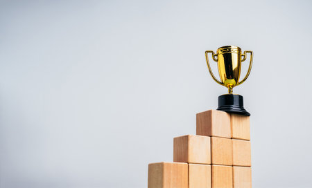 Gold trophy on top of wood cubes as bar graph chart steps on white background with copy space. Business goal and success, achievement, competition, leadership, winner occasion, career growth concepts.の写真素材