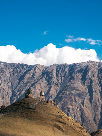 The vertical photo of autumn landscape with huge mountains and blue sky with historical, Gergeti Trinity Church and people on hill near Stepantsminda, on top of mountain in Kazbegi, Georgia.の写真素材