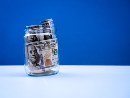 100 dollars bill banknotes, money cash inside glass jar isolated on white table and blue background with copy space. Savings money, future budgets, monthly instalments, business financial concept.の写真素材