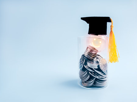 Shining smile face on clear light bulb wear graduation hat, on many coins in glass jar on blue background with copy space. Savings money budget for education, knowledge course, scholarship concept.の写真素材