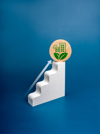 Green growth, ESG business concept. Arrow rise up on white block step graph object with buildings, sun and leaves as sustainable environmentally friendly living icon on wood, blue vertical background.の写真素材