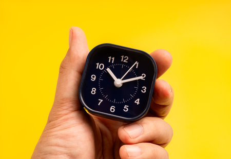 Time management, deadline, punctuality, time tracking, and efficient daily planning, scheduling control concept. Close-up of hand holding black small clock 10:00 on vibrant yellow background.の写真素材