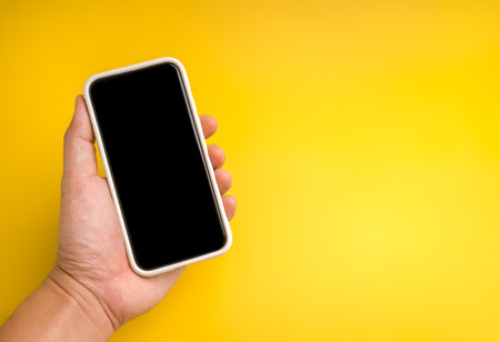 Hand holding smartphone with blank black screen on yellow background, perfect for app design, UI...