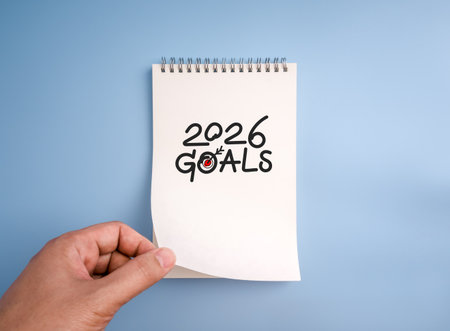 2026 Goals text with hand-drawn target icon on white notebook page held by hand, blue background, representing New Year resolution, goal setting, business planning, and personal development concept.の写真素材