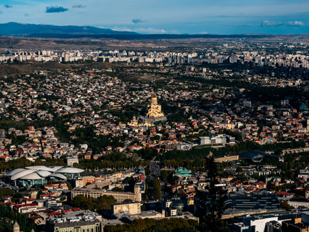 Aerial view of Tbilisi city, Georgia, showcasing urban landscape with Holy Trinity Cathedral at the center, residential buildings, modern architecture, and distant mountain backdrop.の写真素材