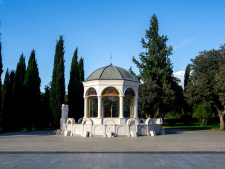 Gazebo-style pavilion inside the Holy Trinity Cathedral grounds in Tbilisi, Georgia, featuring stained glass windows, white columns, and peaceful surroundings with evergreen trees under blue sky.の写真素材