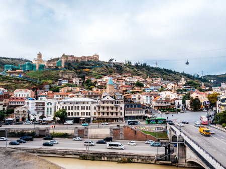 Panoramic cityscape of Tbilisi, Georgia, featuring Old Town buildings, Narikala Fortress on the hill, cable cars, historic churches, and modern traffic along the Kura River under cloudy skies.の写真素材