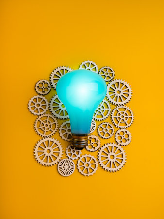 Blue light bulb on top of wooden gear wheels over vertical yellow background, symbolizing idea management, energy systems, workflow innovation, engineering solutions and business process optimization.の写真素材