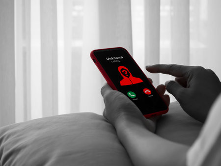 Scammer or fraud call alert concept with unknown caller appearing, red hooded figure on smartphone screen in hands, black and white photo. Phone phishing, cybercrime, and mobile security risk.の写真素材