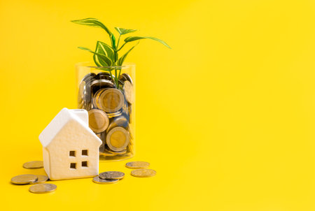 White ceramic house with glass jar full of coins and plant growing inside. Concept of home savings, financial planning, affordable housing, sustainable investment, and long-term wealth growth.の写真素材