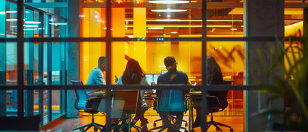 A group of people are seated at a rectangular table, positioned in front of a large window. They appear engaged in conversation, possibly during a meeting or social gathering. The window reveals a view of the outdoors, with natural light streaming into the room.の素材