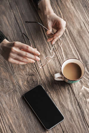 On a wooden table, a cup of coffee, a smartphone and glasses in young female hands. Morning coffee. Copy spaceの写真素材