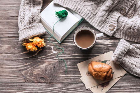 Top view of a cup of coffee on a wooden table and a warm sweater, an open book and a croissant....
