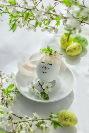 Happy easter. Natural egg with painted cute face in floral wreaths on linen fabric with blooming spring branch, petals and green leaves in soft white light. Creative Easter background.の写真素材