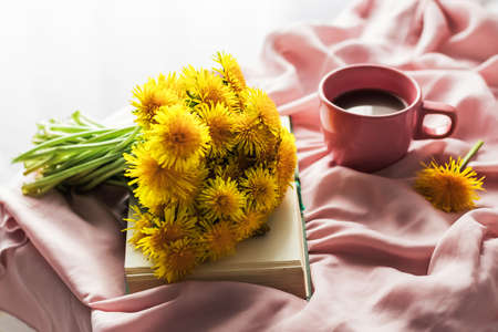 The atmosphere of a romantic morning, coffee in bed. A bouquet of yellow dandelions on an open book and a cup of coffee on a pink silk bed. Home interior. Lifestyle, selective focus.の写真素材