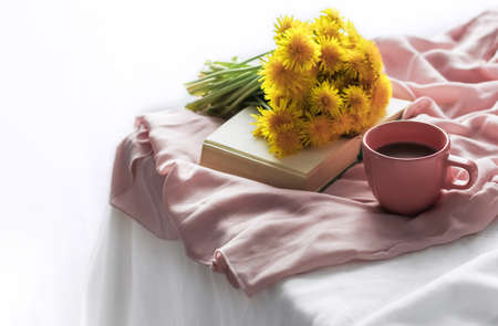 The atmosphere of a romantic morning. A cup of coffee on a table with a pink silk tablecloth, a small bouquet of yellow dandelions on an open book. Home interior. Lifestyle, selective focus, copy spaceの写真素材