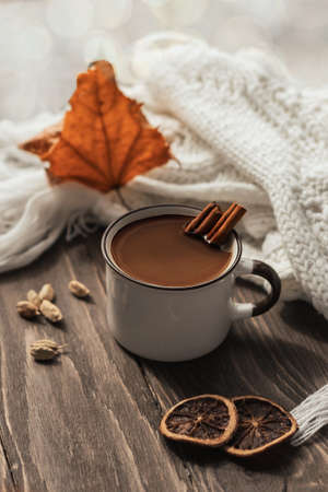 Mug of hot chocolate or cocoa with cinnamon sticks on a wooden table against a background of autumn leaves.Warm scarf and cozy autumn and winter concept.の写真素材