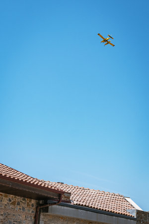 A yellow twin-engine firefighting seaplane flies in a clear blue sky, seen from a low angle above a traditional stone building with a tiled roof in Turkey.の写真素材