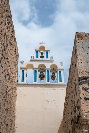 An iconic view of a traditional Greek Orthodox bell tower with three bells and blue accents, framed by rustic stone walls under a cloudy sky in a Santorini village.の写真素材