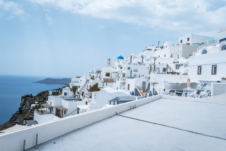A stunning view of the classic whitewashed buildings of a Santorini village stacked on a hillside, with a prominent church featuring a blue dome and a bell tower, and tourists walking along the paths.の写真素材