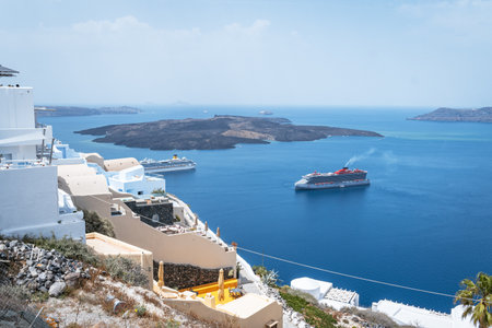 A wide panoramic view of the iconic Santorini caldera with multiple cruise ships in the deep blue sea, surrounding the volcanic island of Nea Kameni under a hazy summer sky.の写真素材