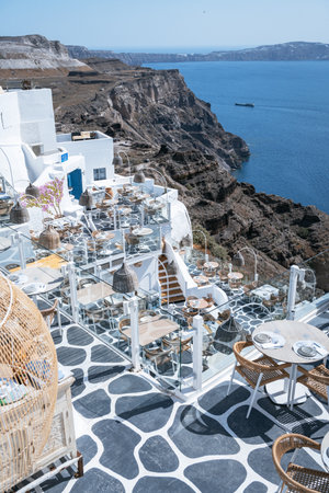 A stunning view of the classic whitewashed buildings of a Santorini village stacked on a hillside, with a prominent church featuring a blue dome and a bell tower, and tourists walking along the paths.の写真素材