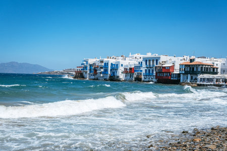 The iconic white and colorful buildings of Little Venice in Mykonos stand right at the water's edge, with waves breaking on a rocky beach.の写真素材