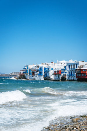 The iconic white and colorful buildings of Little Venice in Mykonos stand right at the water's edge, with waves breaking on a rocky beach.の写真素材