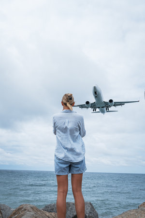 A person stands on a rocky shore, watching a passenger airplane flying low over the sea with its landing gear down.の写真素材