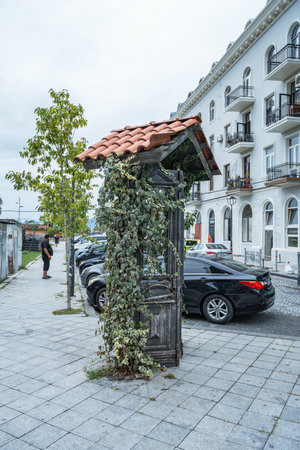 A sunless, old wooden structure with a red tile roof and climbing ivy, standing on a cobblestone sidewalk next to parked cars and a building with balconies.の写真素材
